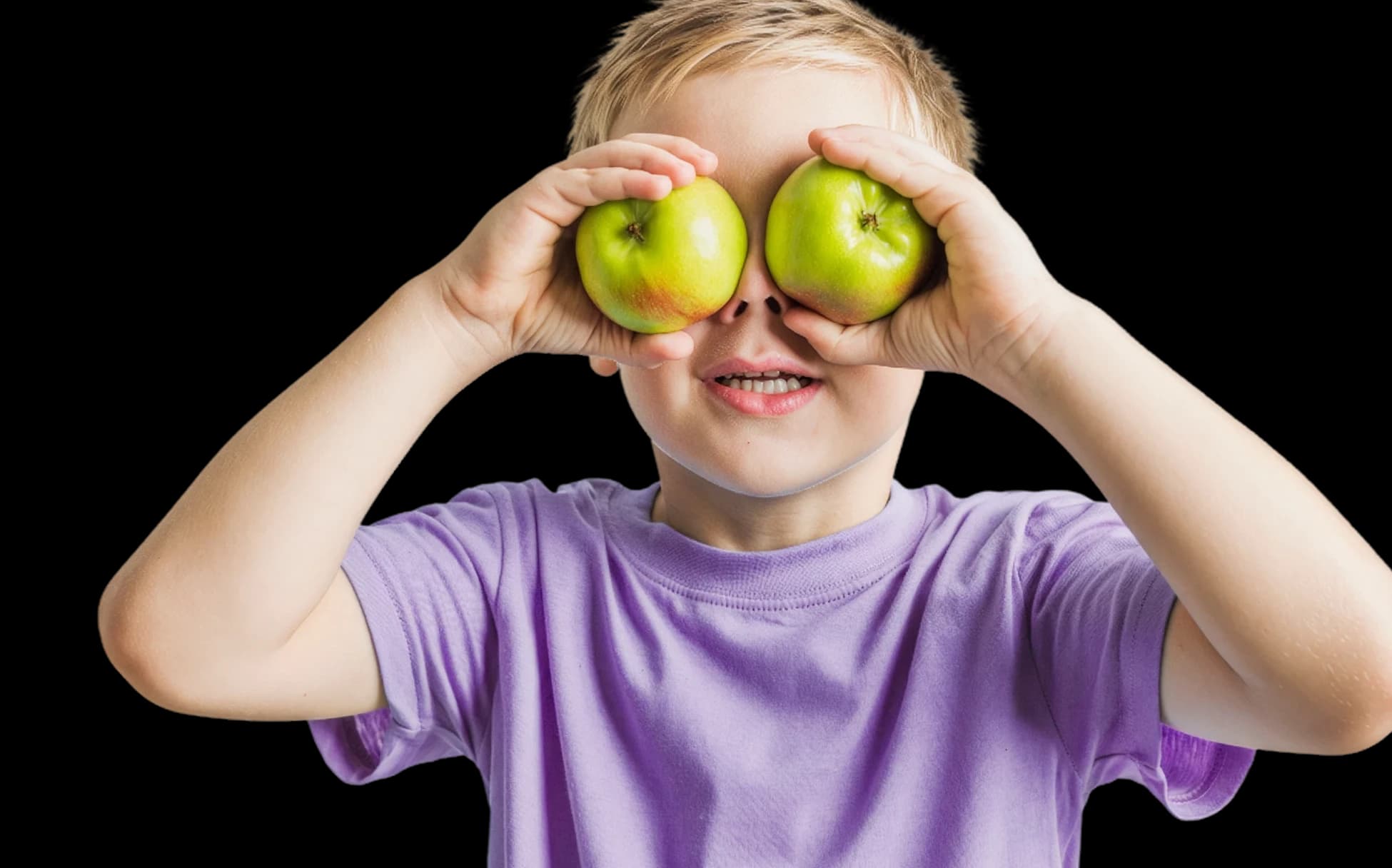 Criança sorrindo com duas maçãs verdes sobre os olhos